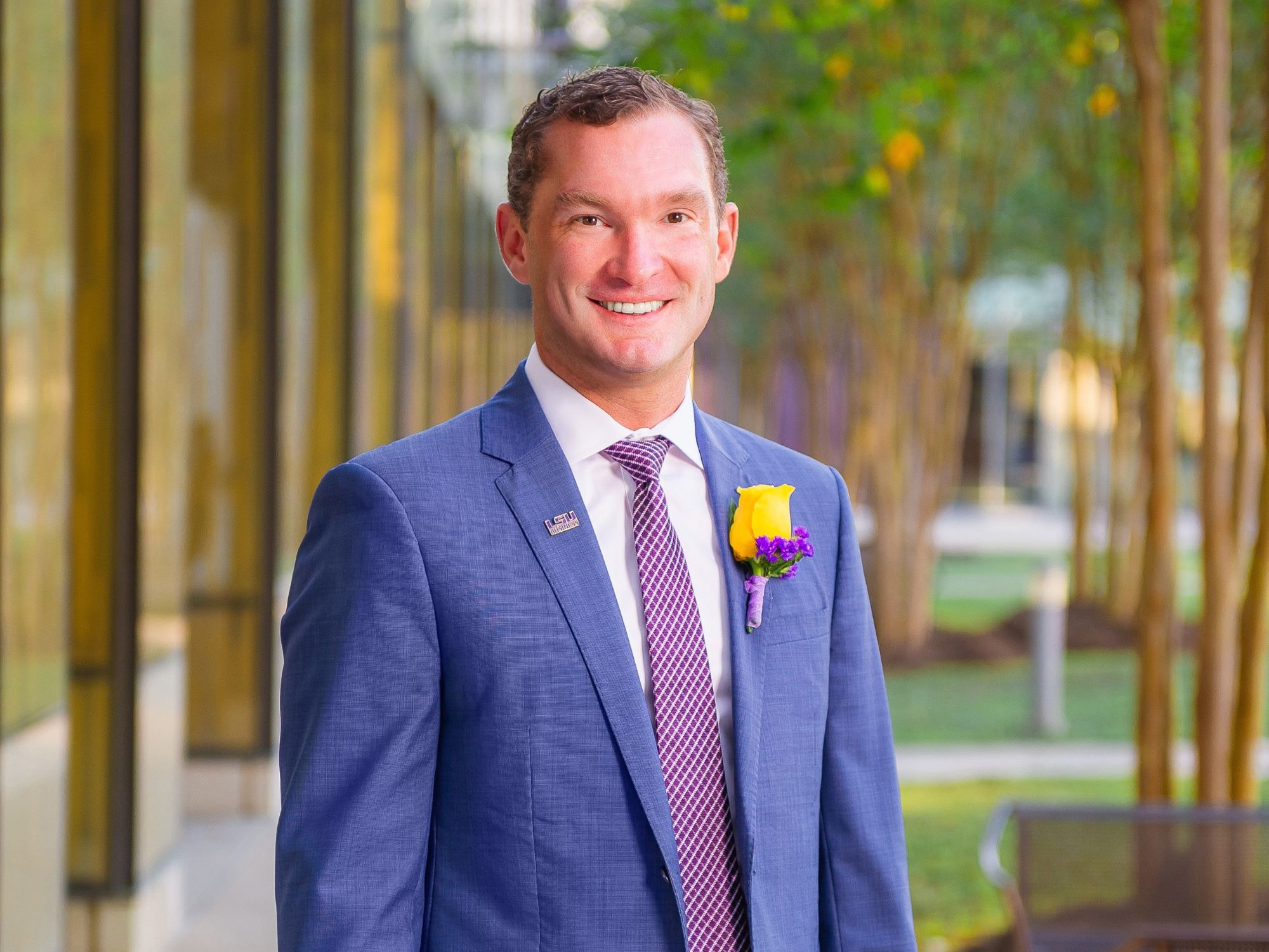 Nick Speyrer in a dark suit. He is standing outside with trees and columns in the background. 