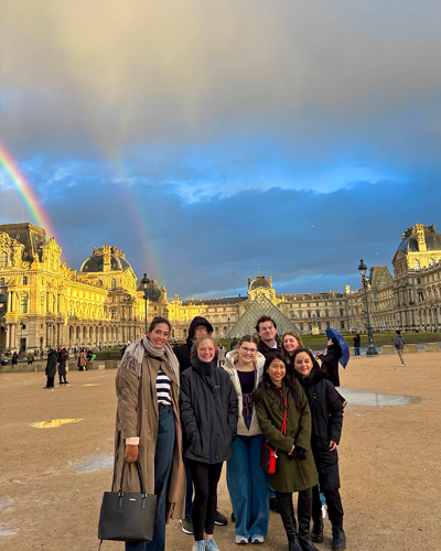 Natalie Wagaman stands with a group of students on a rainy day in Paris. In the background, you can spot double rainbows over the Louvre.