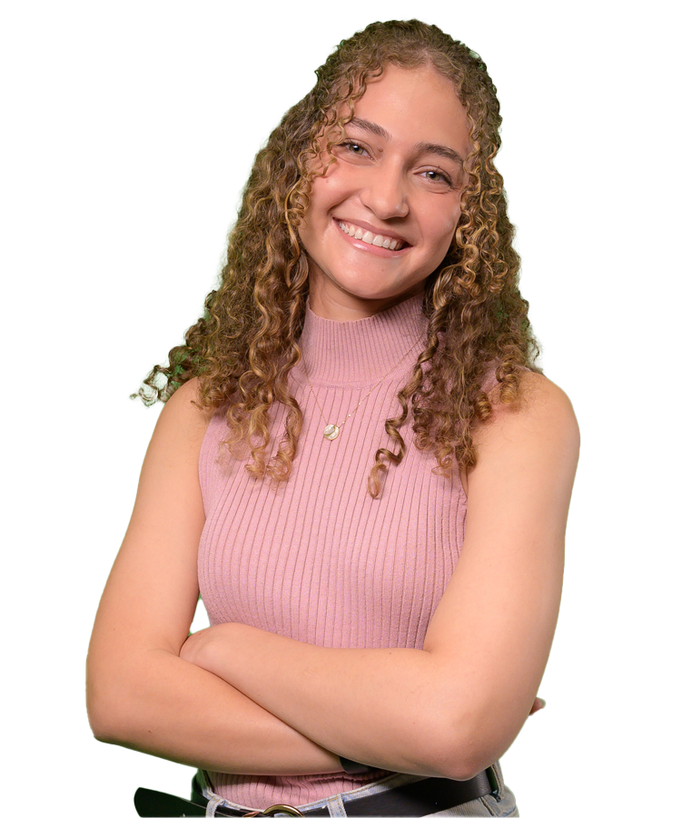 Ranna wears jeans and a pale pink sleeveless sweater. Her arms are crossed in front of her and her head is tilted to the right. She flashes a confident smile.